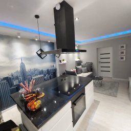 WYKOŃCZENIA WNĘTRZ-GRZEGORZ BATOR - Nowoczesna kuchnia z czarnym blatem i białymi szafkami, na blacie owoce w szklanej misie, w tle fototapeta z panoramą Nowego Jorku i niebieskie oświetlenie LED podsufitowe.