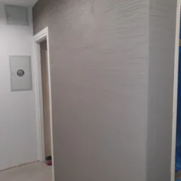 Szara ściana z fakturą imitującą rysunek fal, narożnik wykończony w tym samym stylu, widoczna skrzynka z licznikiem na sąsiedniej białej ścianie oraz fragment ościeżnicy drzwiowej.