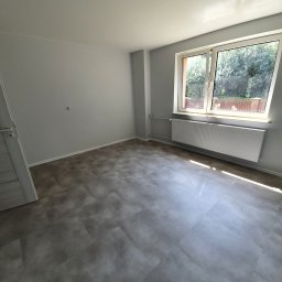 WYKOŃCZENIA WNĘTRZ-GRZEGORZ BATOR - Pusty pokój po remoncie z szarymi ścianami, panelami podłogowymi i grzejnikiem pod oknem, widoczne gniazdko elektryczne na ścianie.