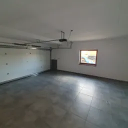 Wykafelkowane wnętrze garażu z bramą segmentową i automatyką, widok na szare płytki podłogowe w formacie kwadratu i okno w tle.