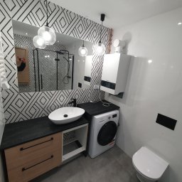 WYKOŃCZENIA WNĘTRZ-GRZEGORZ BATOR - Nowoczesna łazienka z umywalką nablatową, szafką z drewnianymi szufladami, pralką, grzejnikiem drabinkowym i wzorzystymi płytkami na ścianie, oświetlona wiszącymi lampami z kulistymi kloszami.