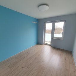 WYKOŃCZENIA WNĘTRZ-GRZEGORZ BATOR - Pomalowane na niebiesko wnętrze pokoju z drewnianą podłogą, białymi drzwiami balkonowymi i szarą ścianą, oświetlone okrągłą lampą sufitową.