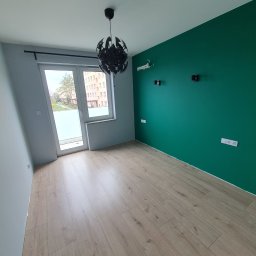 WYKOŃCZENIA WNĘTRZ-GRZEGORZ BATOR - Pomalowany na zielono pokój z drewnianą podłogą, widoczne gniazdka elektryczne, lampa sufitowa i okno balkonowe z widokiem na budynek mieszkalny.