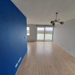 WYKOŃCZENIA WNĘTRZ-GRZEGORZ BATOR - Pomalowane na biało i granatowo wnętrze mieszkania z drewnianą podłogą, widok na balkon i nowoczesną lampę sufitową.