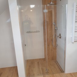 WYKOŃCZENIA WNĘTRZ-GRZEGORZ BATOR - Kabina prysznicowa ze szklanymi drzwiami, wykończona białymi i drewnopodobnymi płytkami, z widocznym odpływem liniowym w podłodze i odbiciem fotografa.
