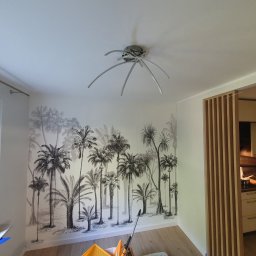 WYKOŃCZENIA WNĘTRZ-GRZEGORZ BATOR - Pokój po dekoracyjnym malowaniu ścian z motywem tropikalnych palm, widoczna nowoczesna lampa sufitowa i fragment kuchni za drewnianą ścianką działową.