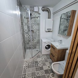 WYKOŃCZENIA WNĘTRZ-GRZEGORZ BATOR - Wykończona łazienka w bieli i szarości: kabina prysznicowa, pralka, umywalka z szafką z drewnianymi akcentami, mozaika na podłodze i części ścian.