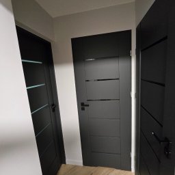 WYKOŃCZENIA WNĘTRZ-GRZEGORZ BATOR - Nowoczesne, czarne drzwi wewnętrzne z poziomymi frezowaniami i klamkami na tle jasnych ścian i parkietu w jodełkę. Widoczne trzy skrzydła drzwiowe.