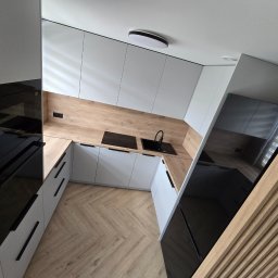 WYKOŃCZENIA WNĘTRZ-GRZEGORZ BATOR - Nowoczesna kuchnia w bieli i drewnie: zabudowa z czarnymi akcentami, parkiet w jodełkę i oświetlenie sufitowe. Ujęcie z góry, podkreślające układ i przestrzeń.