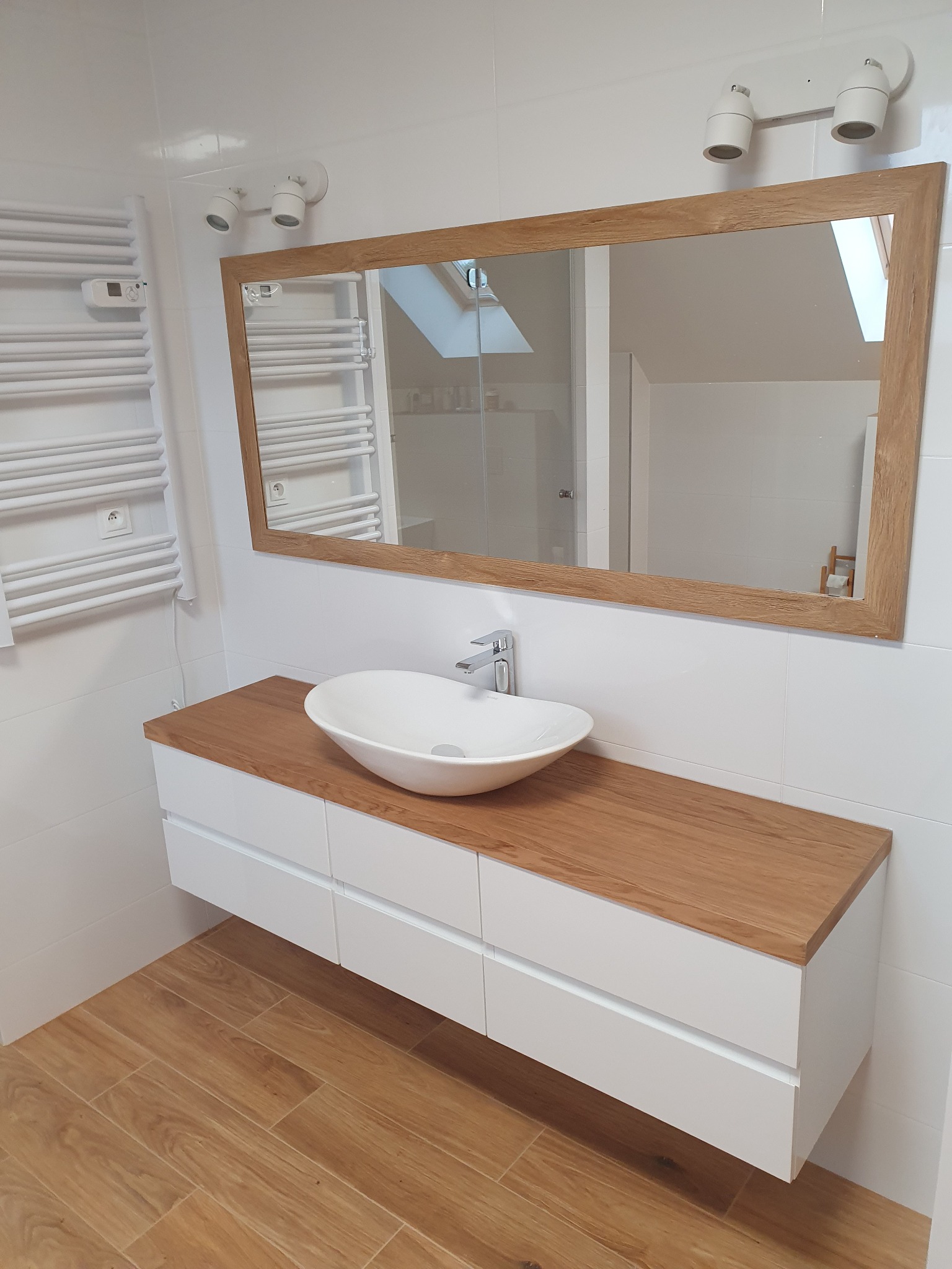 Nowoczesna łazienka z białymi płytkami, drewnianą podłogą i blatem pod umywalkę, owalna umywalka nablatowa i lustro w drewnianej ramie odbijające skos dachu.