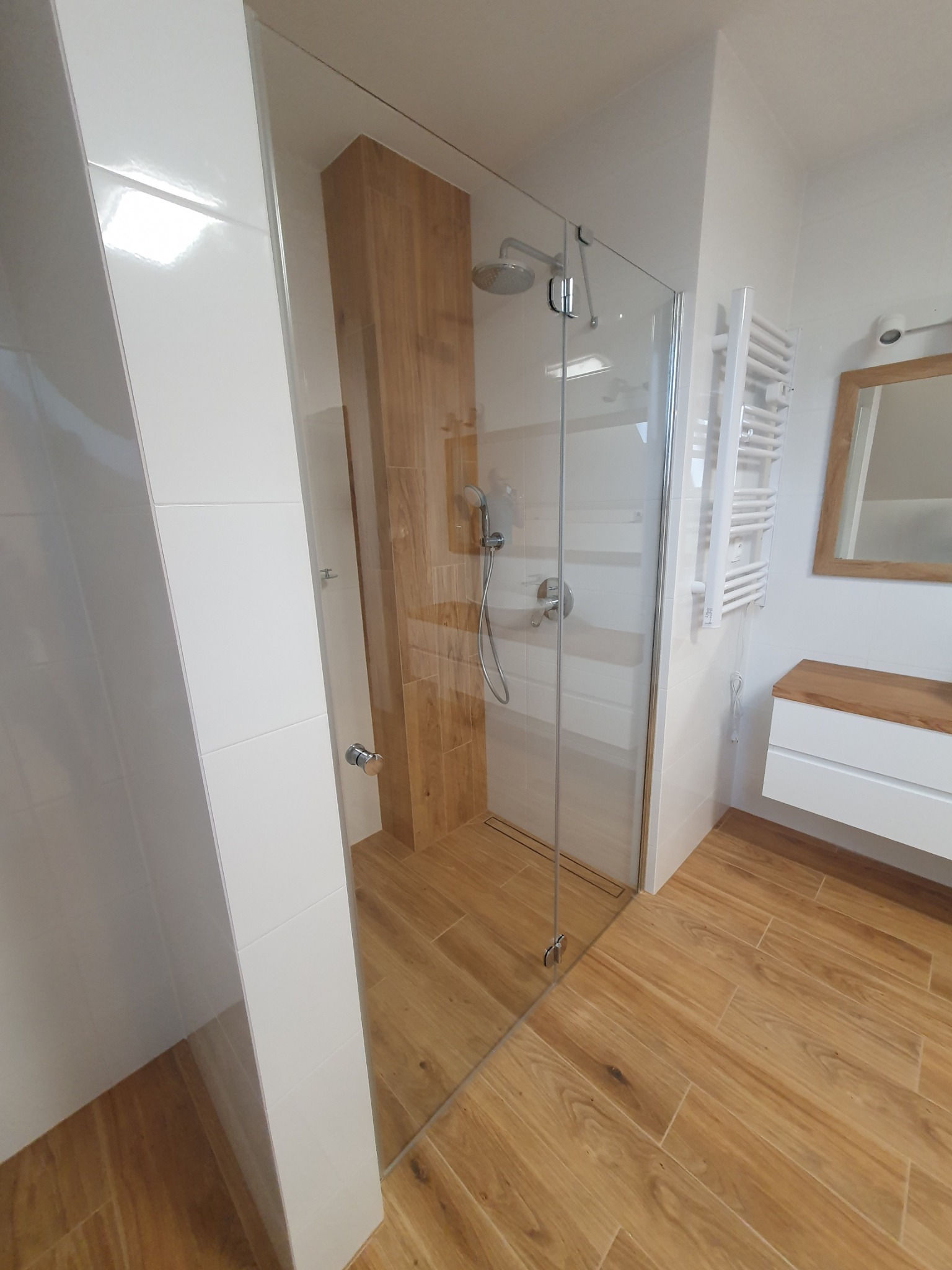 Nowoczesna łazienka z przeszkloną kabiną prysznicową typu walk-in, drewnopodobne płytki na podłodze i ścianie prysznica, białe płytki na pozostałych ścianach, grzejnik drabinkowy i szafka...