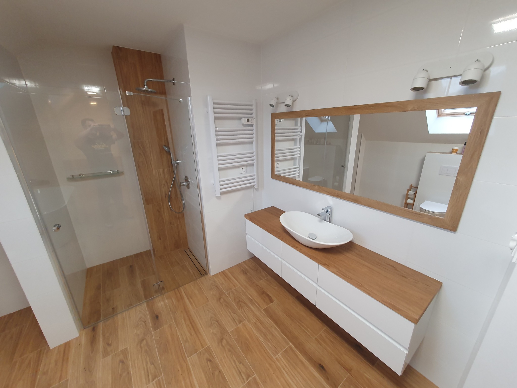 Nowoczesna łazienka z kabiną prysznicową z drewnianym wykończeniem, białą umywalką nablatową na szafce z drewnianym blatem i dużym lustrem w drewnianej ramie.