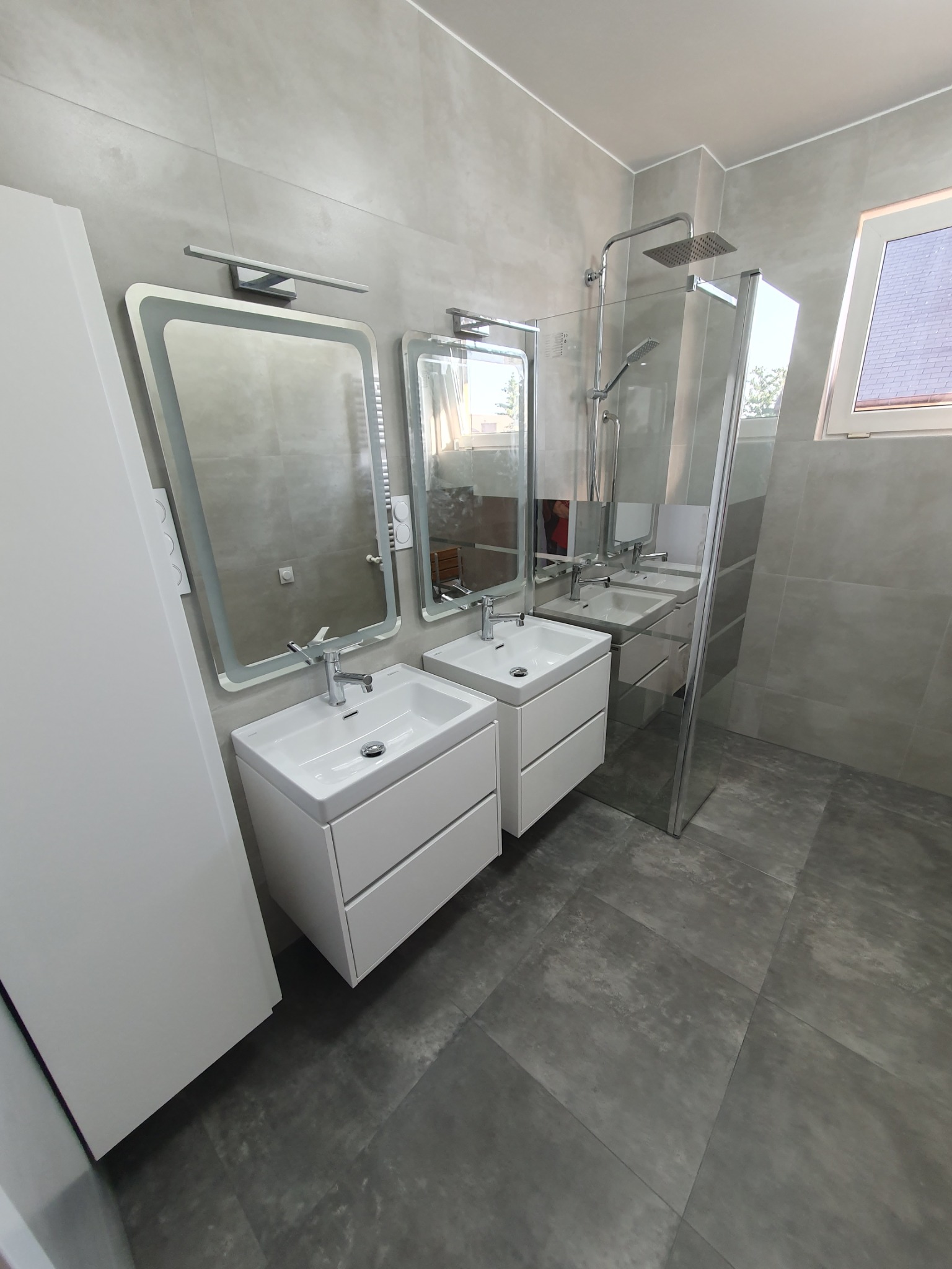 Nowoczesna łazienka w odcieniach szarości z dwoma umywalkami nablatowymi na białych szafkach, podświetlanymi lustrami i kabiną prysznicową typu walk-in z deszczownicą.