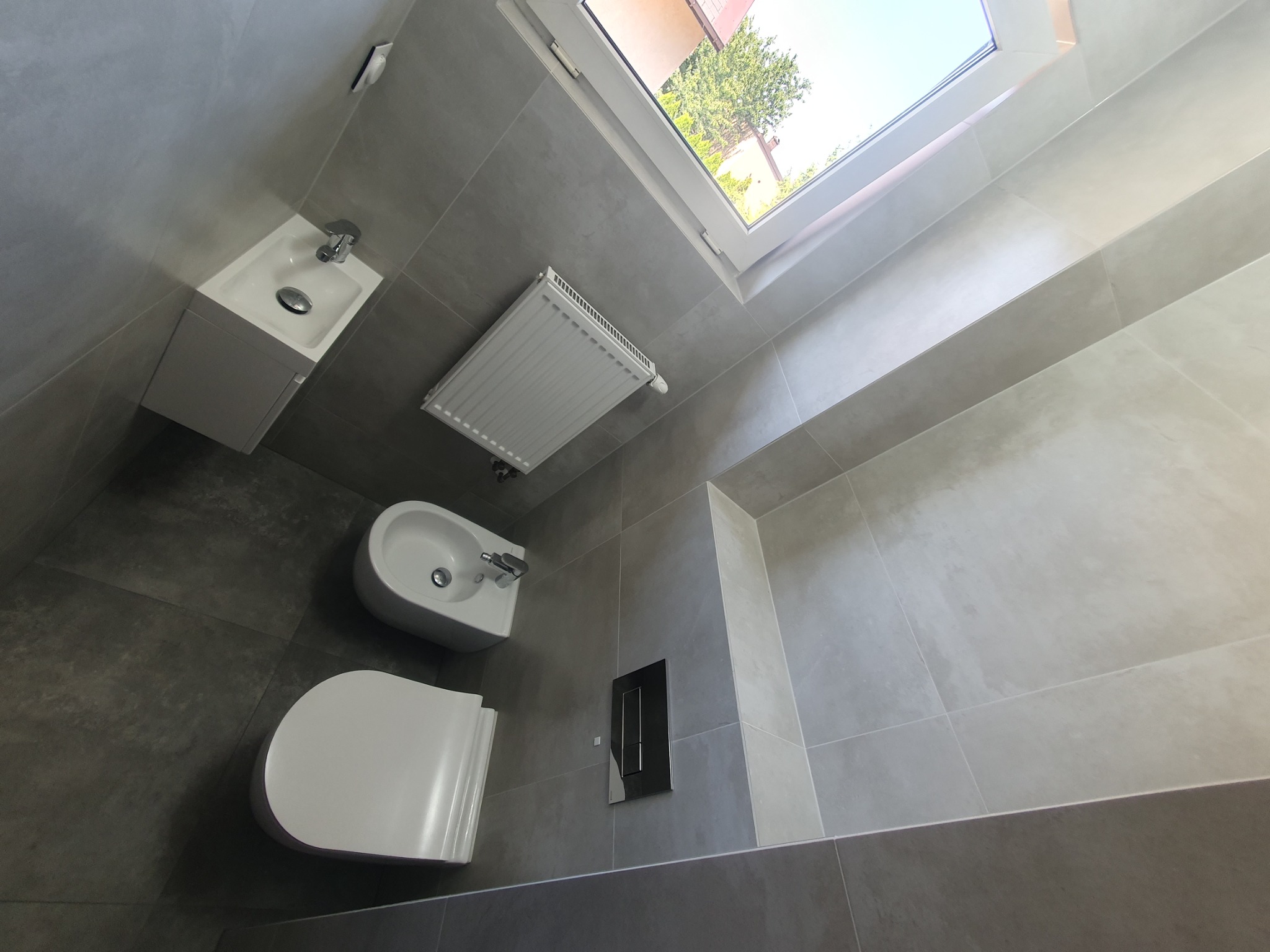 Wykończona łazienka w szarościach z białym wyposażeniem: wisząca toaleta, bidet i umywalka z szafką, grzejnik drabinkowy pod oknem dachowym.