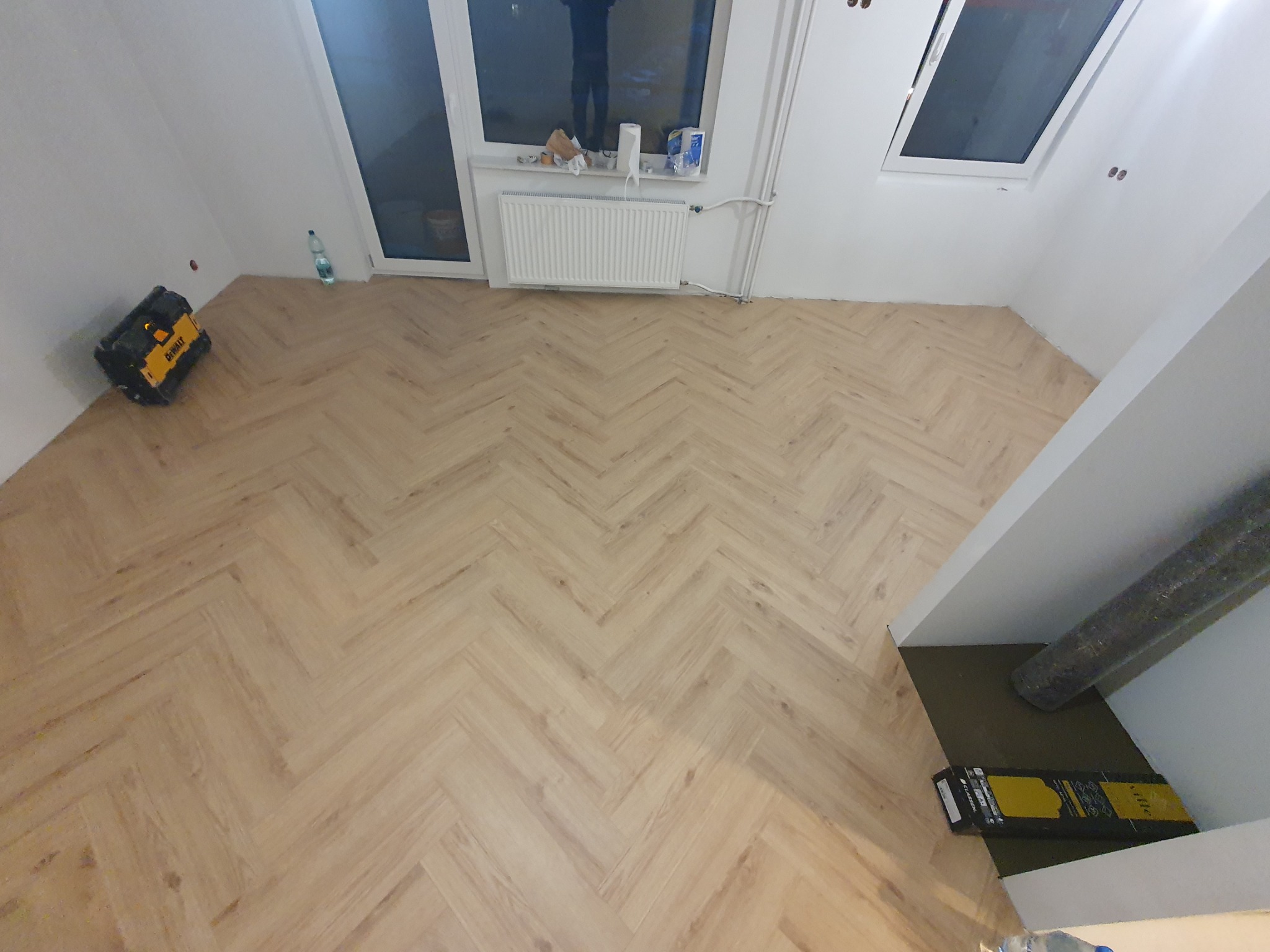 Ukończona podłoga z paneli ułożonych w jodełkę w jasnym pomieszczeniu z oknami dachowymi i grzejnikiem, widoczna butelka wody i skrzynka narzędziowa DeWalt.