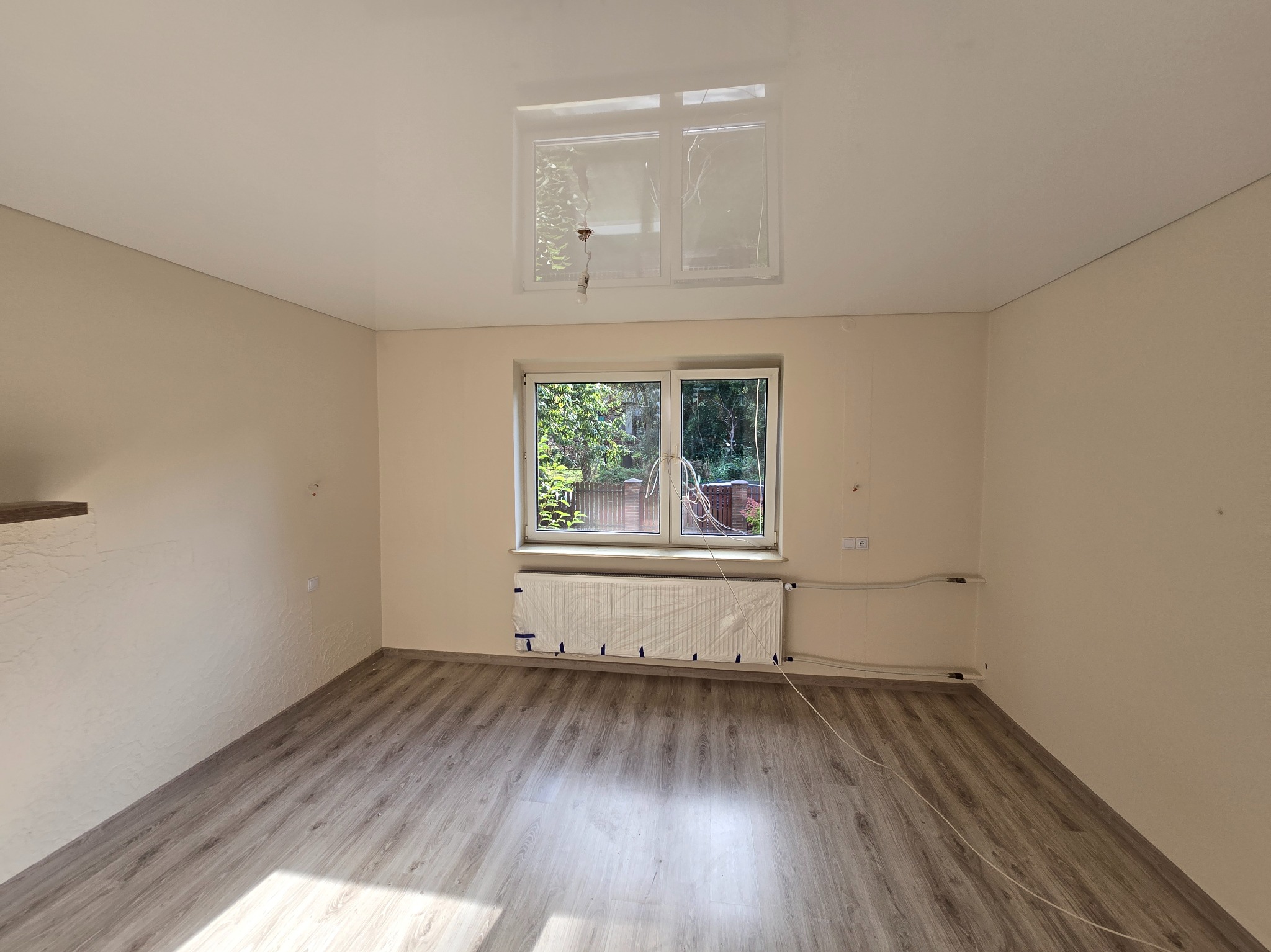 Jasne wnętrze po remoncie w Będzinie: podłoga z paneli, świeżo pomalowane ściany w odcieniach beżu, okno z widokiem na zieleń, zabezpieczony grzejnik.