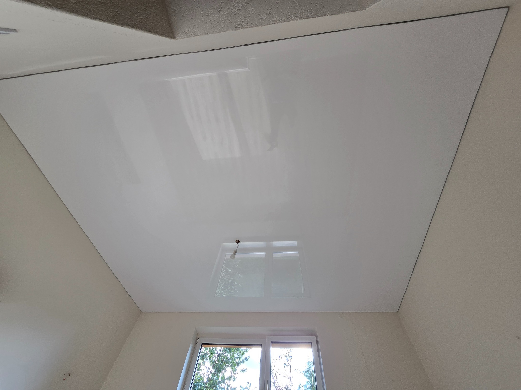 Biały, błyszczący sufit napinany w pokoju z oknem. Widoczne odbicie okna w suficie. Minimalistyczne wykończenie.