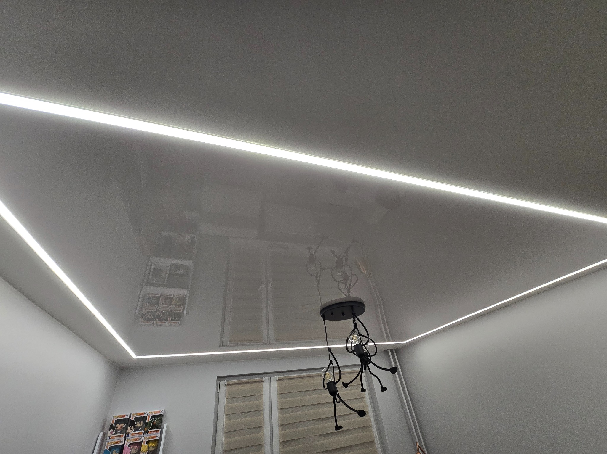 Biały, podwieszany sufit z połyskiem i oświetleniem LED wzdłuż krawędzi. W tle widoczna lampa i okno z żaluzjami.