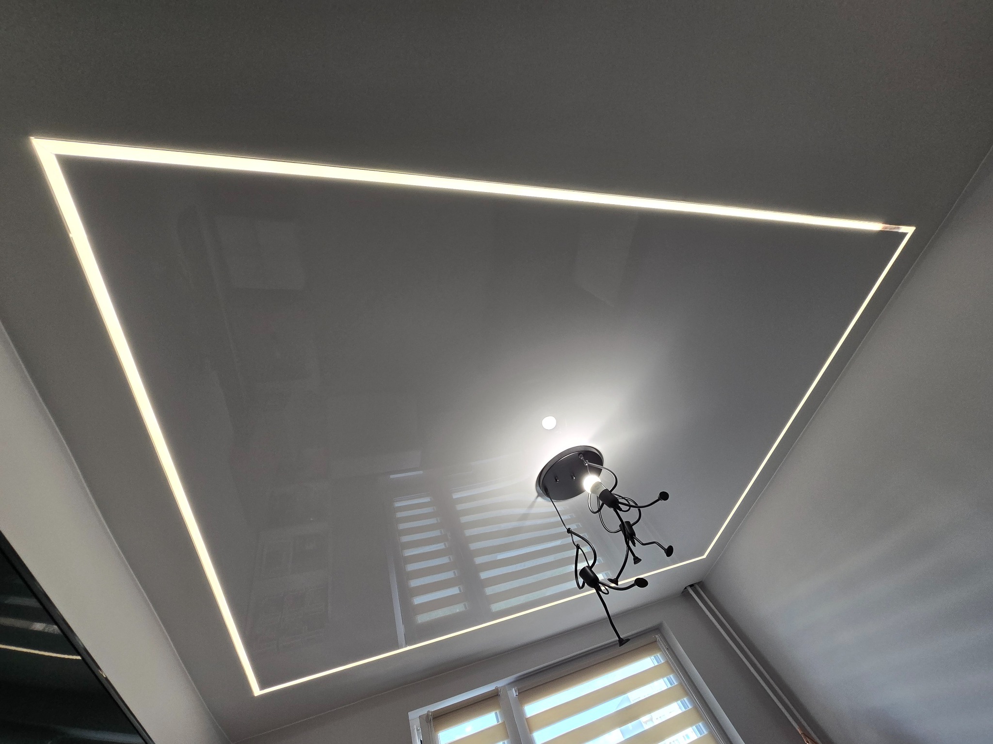 Biały, błyszczący sufit napinany z wbudowanym oświetleniem LED w kształcie prostokąta. W tle widoczna lampa i okno z żaluzjami.
