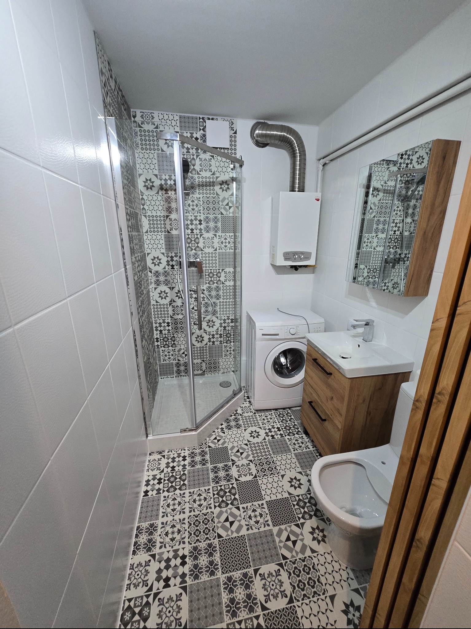 Wykończona łazienka w bieli i szarości: kabina prysznicowa, pralka, umywalka z szafką z drewnianymi akcentami, mozaika na podłodze i części ścian.