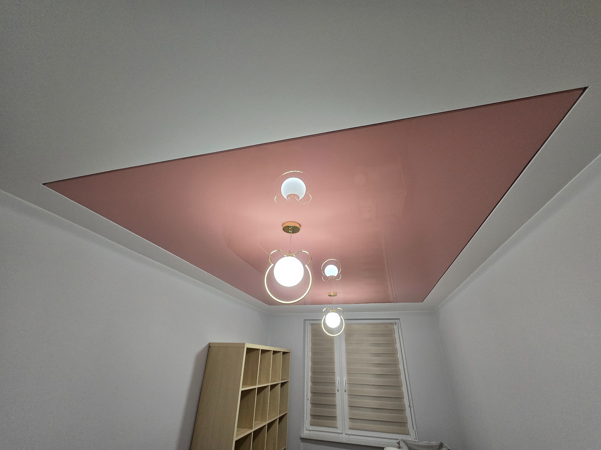 Nowoczesny sufit podwieszany z różowym panelem i designerskimi lampami w kształcie misia, widok z dołu, jasne wnętrze pokoju z oknem i regałem.