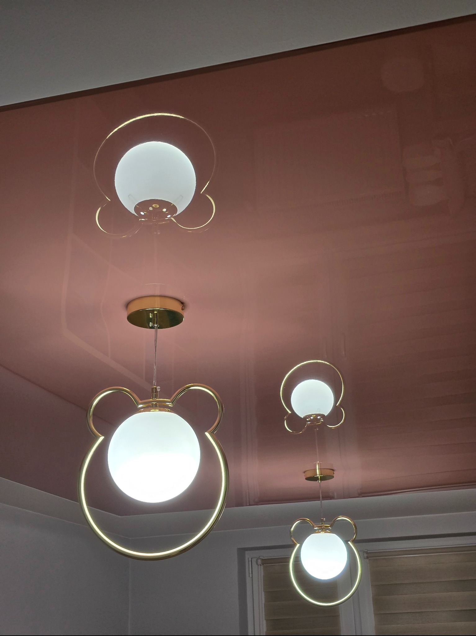 Błyszczący, różowy sufit z zamontowanymi lampami w kształcie głowy misia, z okrągłymi, mlecznymi kloszami i złotymi obwódkami. Widoczne odbicia w suficie.