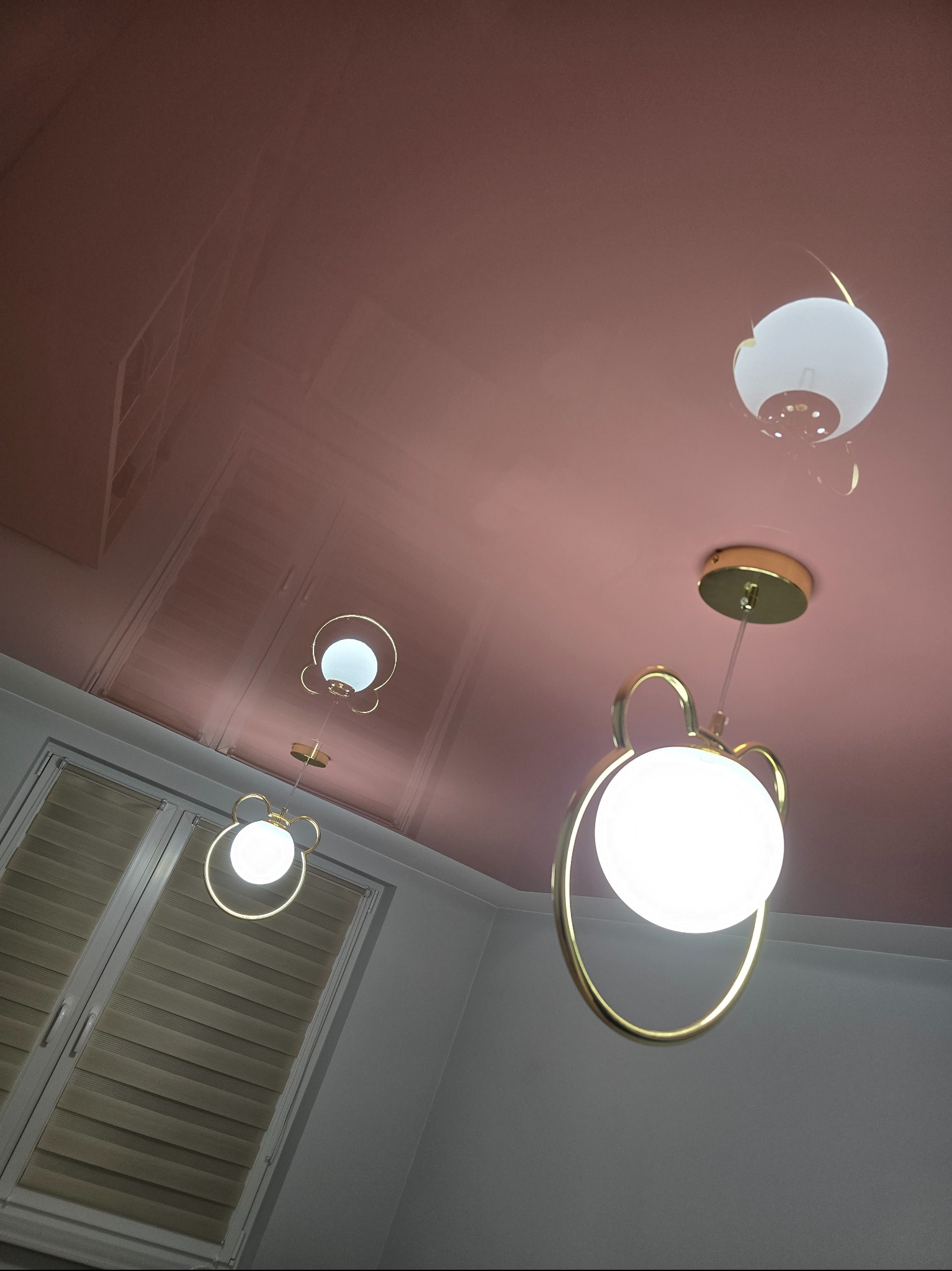 Białe, kuliste lampy z złotymi elementami na tle błyszczącego, różowego sufitu i okna z żaluzjami. Ujęcie z dołu, podkreślające nowoczesny design oświetlenia.