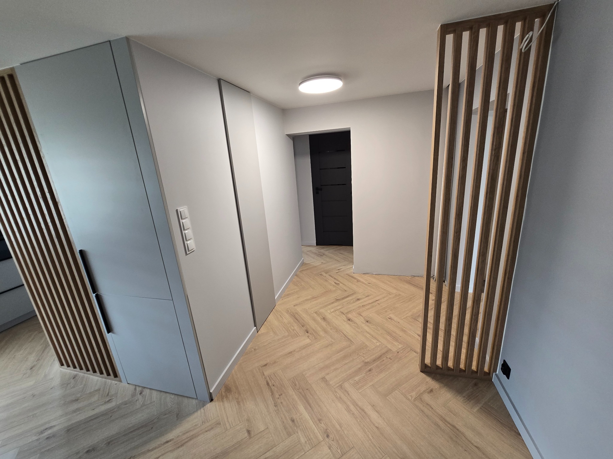 Nowoczesny przedpokój z szafą w zabudowie i drewnianą ażurową ścianką działową. Parkiet ułożony w jodełkę. Minimalistyczne oświetlenie sufitowe.