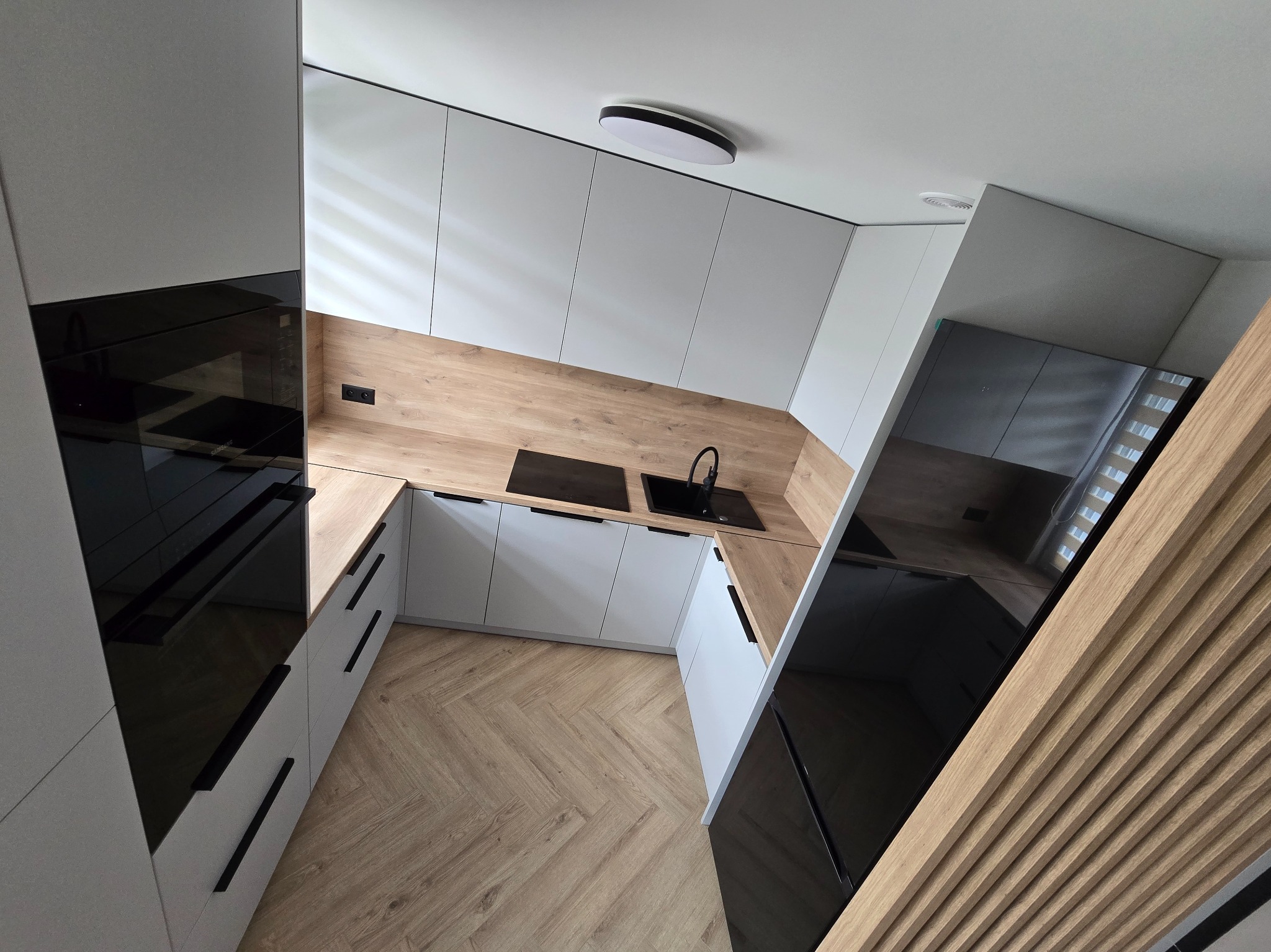 Nowoczesna kuchnia w bieli i drewnie: zabudowa z czarnymi akcentami, parkiet w jodełkę i oświetlenie sufitowe. Ujęcie z góry, podkreślające układ i przestrzeń.