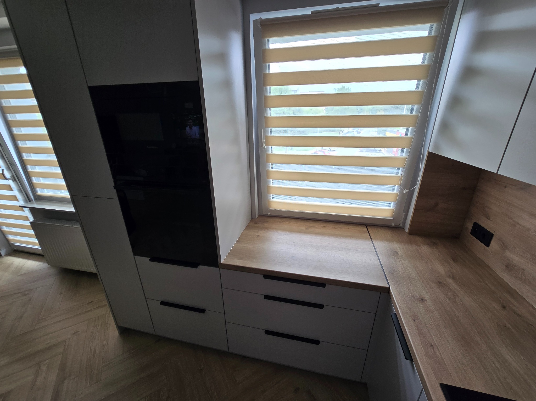 Nowoczesna zabudowa kuchenna z białymi szafkami i drewnianym blatem, zintegrowana z oknem z roletami dzień-noc i parkietem w jodełkę na podłodze.