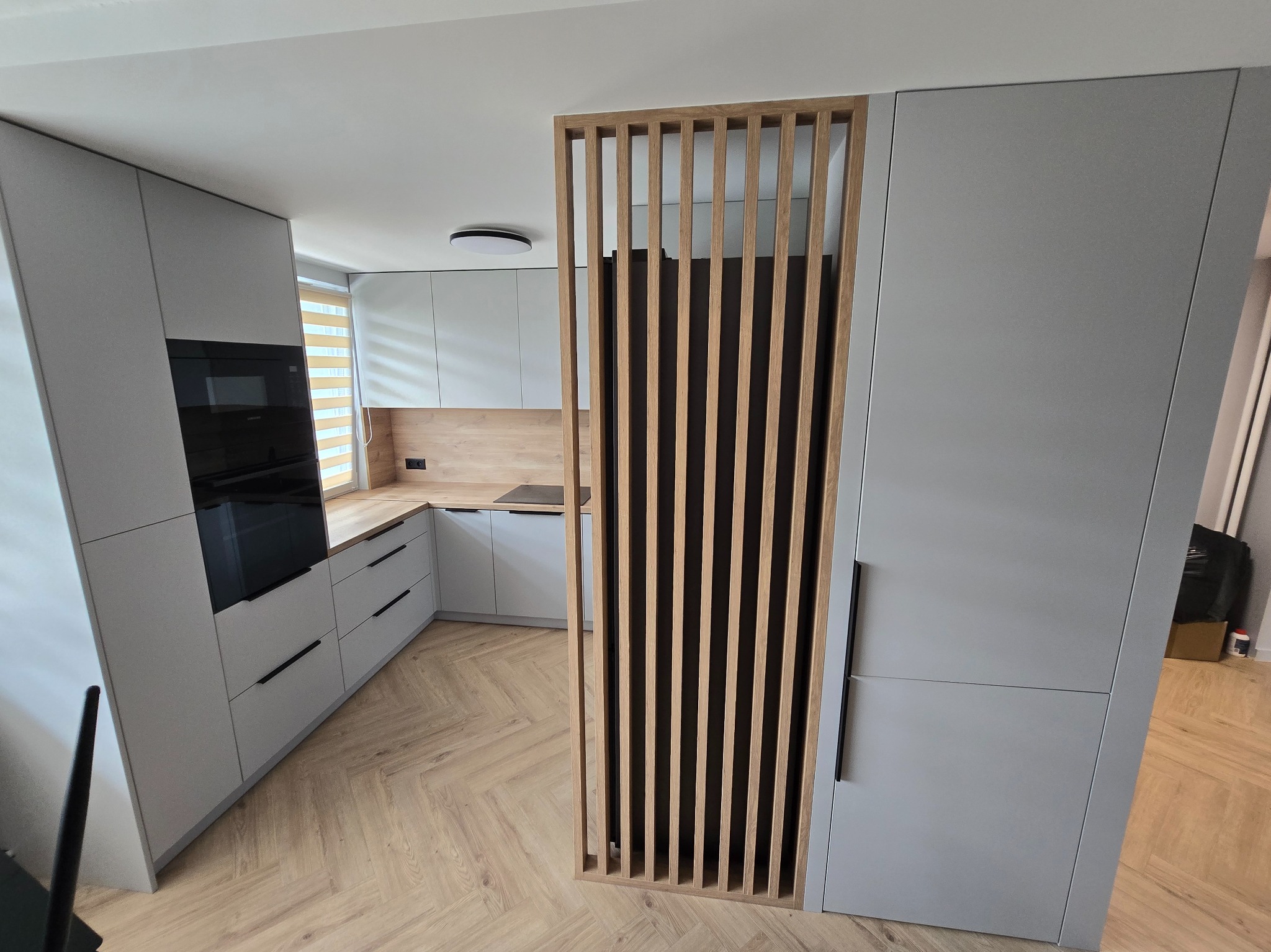 Nowoczesna kuchnia w szarościach z drewnianym panelem ażurowym, parkiet w jodełkę. Minimalistyczny design, zabudowa kuchenna.