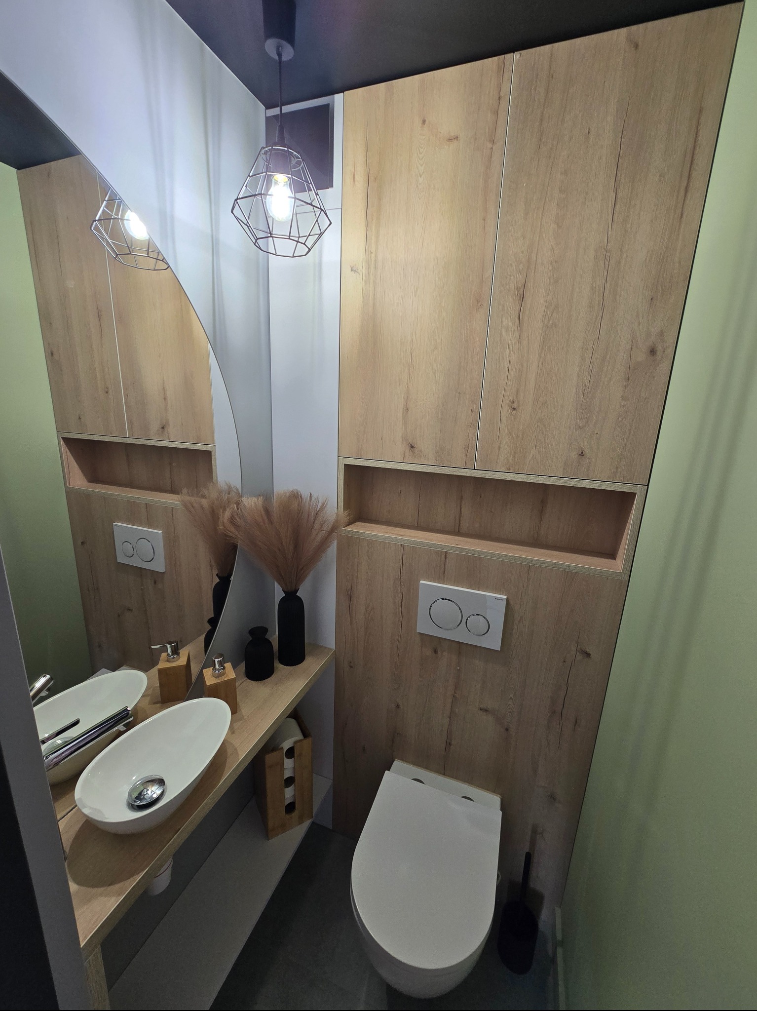 Nowoczesna łazienka z drewnianymi szafkami, asymetrycznym lustrem i owalną umywalką. Minimalistyczny design z akcentami dekoracyjnymi, takimi jak lampa i wazon.