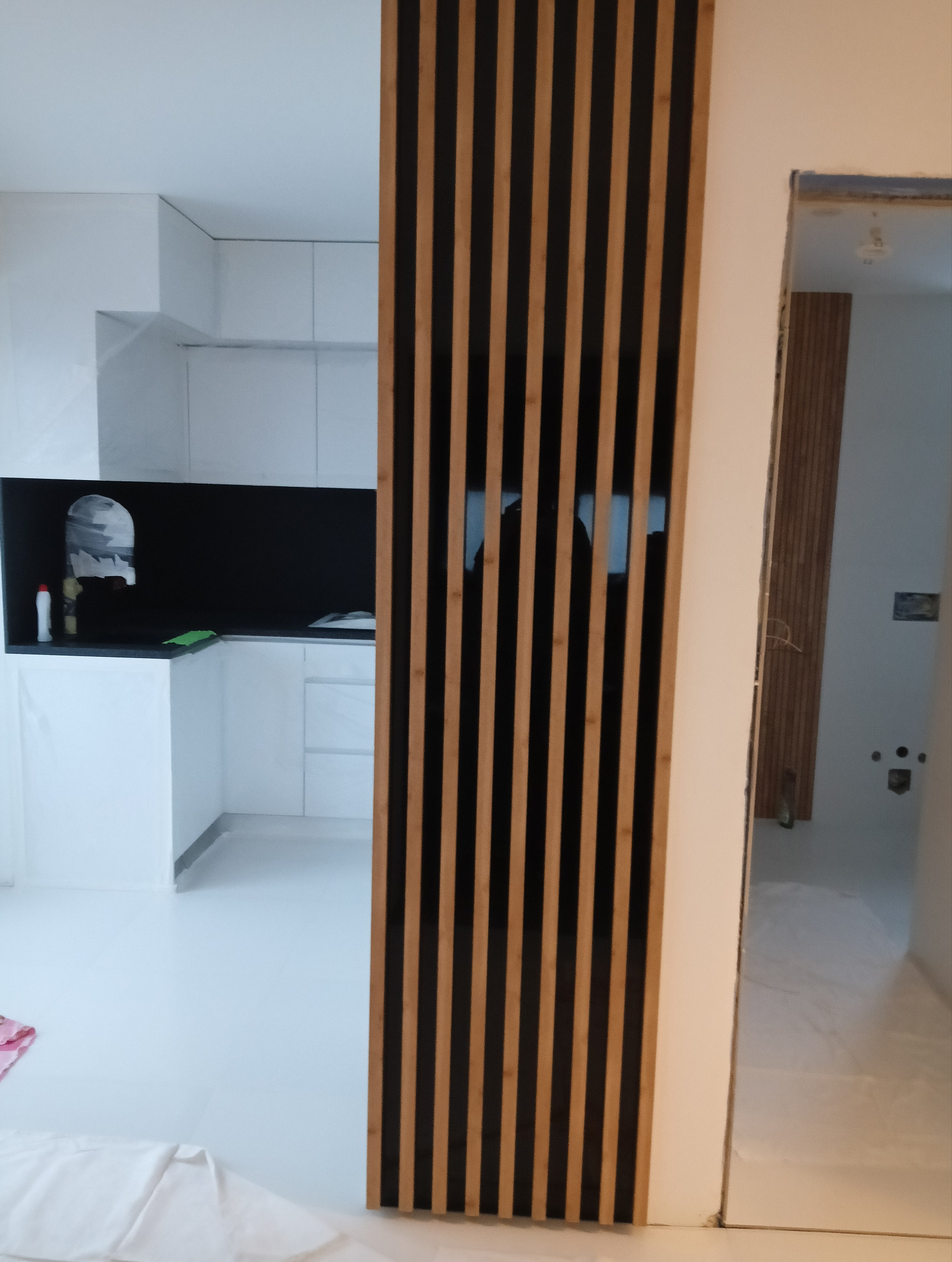 Nowoczesna kuchnia z białymi szafkami i czarnym panelem nad blatem, oddzielona od innego pomieszczenia dekoracyjną ścianką z drewnianych listew w kolorze jasnego drewna i czerni, widok...