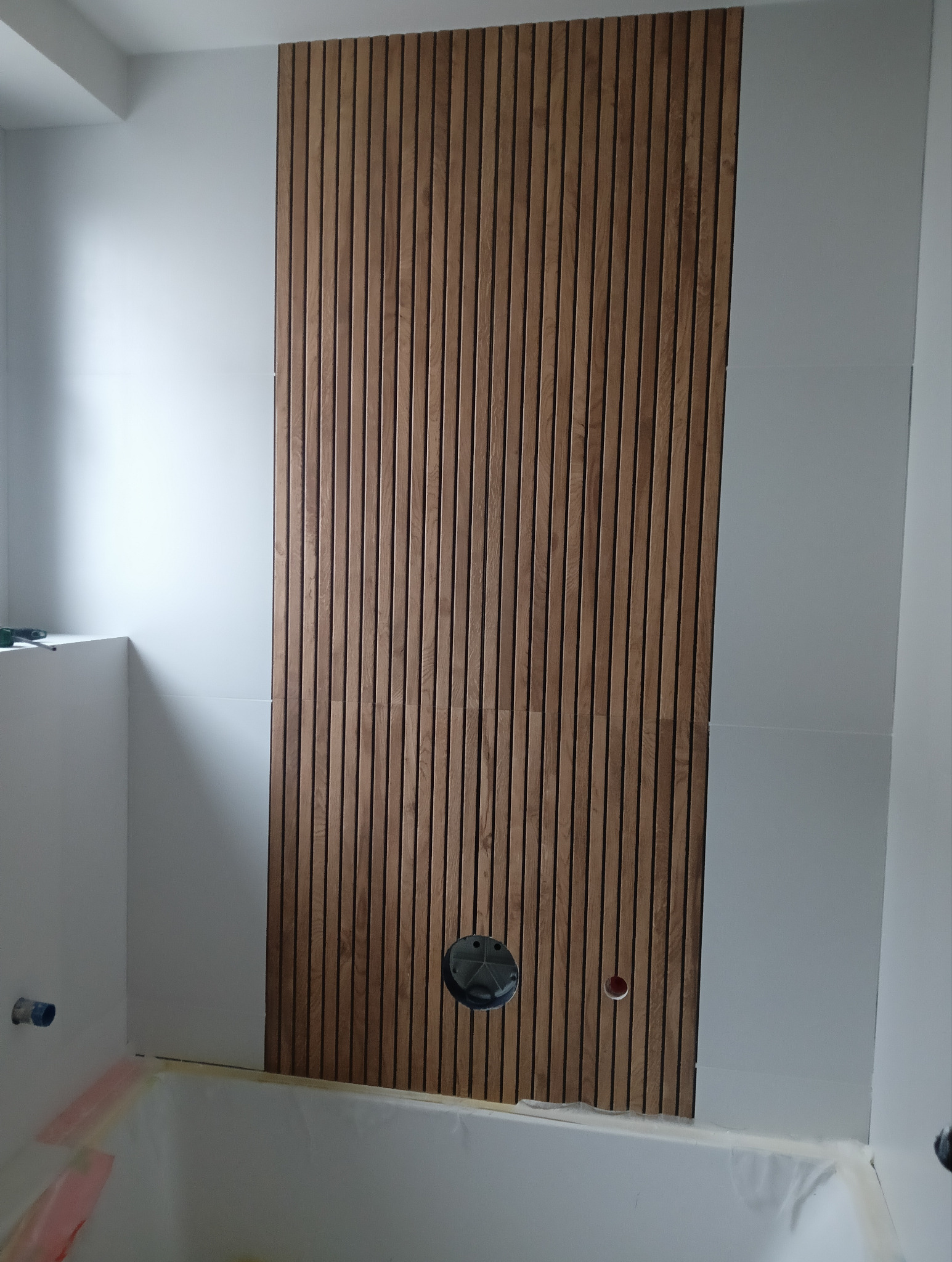 Ściana w łazience z drewnianymi panelami pionowymi i otworem na instalację hydrauliczną w trakcie montażu.