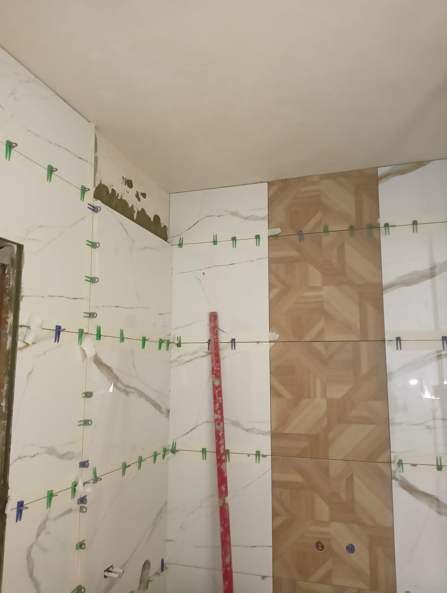 Układanie płytek na ścianie w łazience, widoczne płytki imitujące marmur i drewno, poziomica oraz system poziomowania płytek z klipsami.