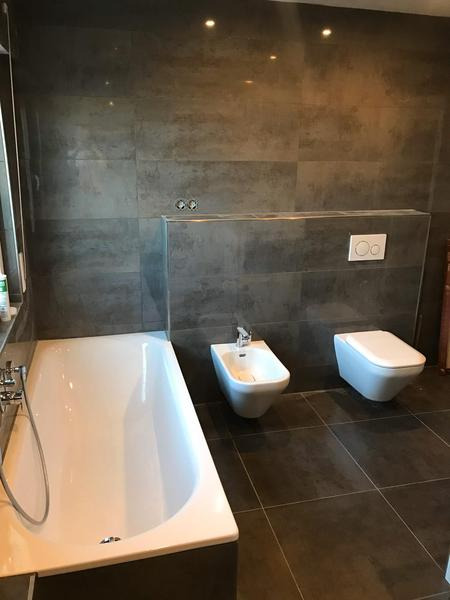 Nowoczesna łazienka z wanną, bidetem i podwieszaną toaletą, wykończona szarą płytką imitującą beton, z zamontowanym panelem sterującym spłuczką.