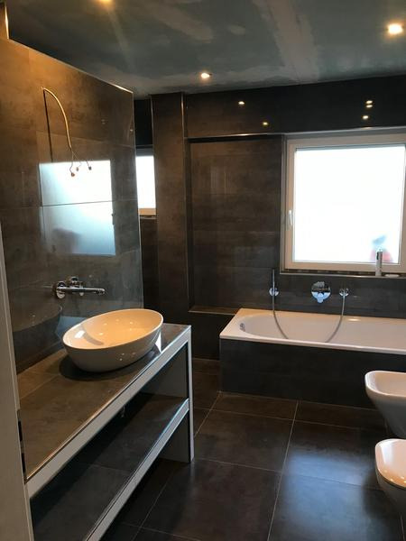 Nowoczesna łazienka z ciemnymi płytkami, wolnostojącą umywalką na metalowym stelażu, wanną i bidetem; sufit przygotowany do malowania.