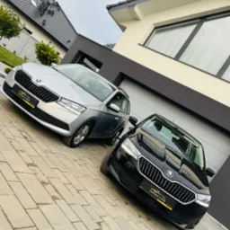 Dwa samochody Skoda, srebrny i czarny, stoją zaparkowane na podjeździe przed nowoczesnym domem z szarą bramą garażową.