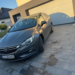 PLATINIUM ERGOLINE MATEUSZ GMEREK - Szary samochód kombi marki Opel Astra zaparkowany na posesji przed nowoczesnym domem z garażem, widoczne ślady wilgoci na kostce brukowej.