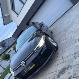 PLATINIUM ERGOLINE MATEUSZ GMEREK - Czarny samochód osobowy marki Volkswagen zaparkowany na kostce brukowej przed nowoczesnym domem z szarą bramą garażową i dużym oknem.