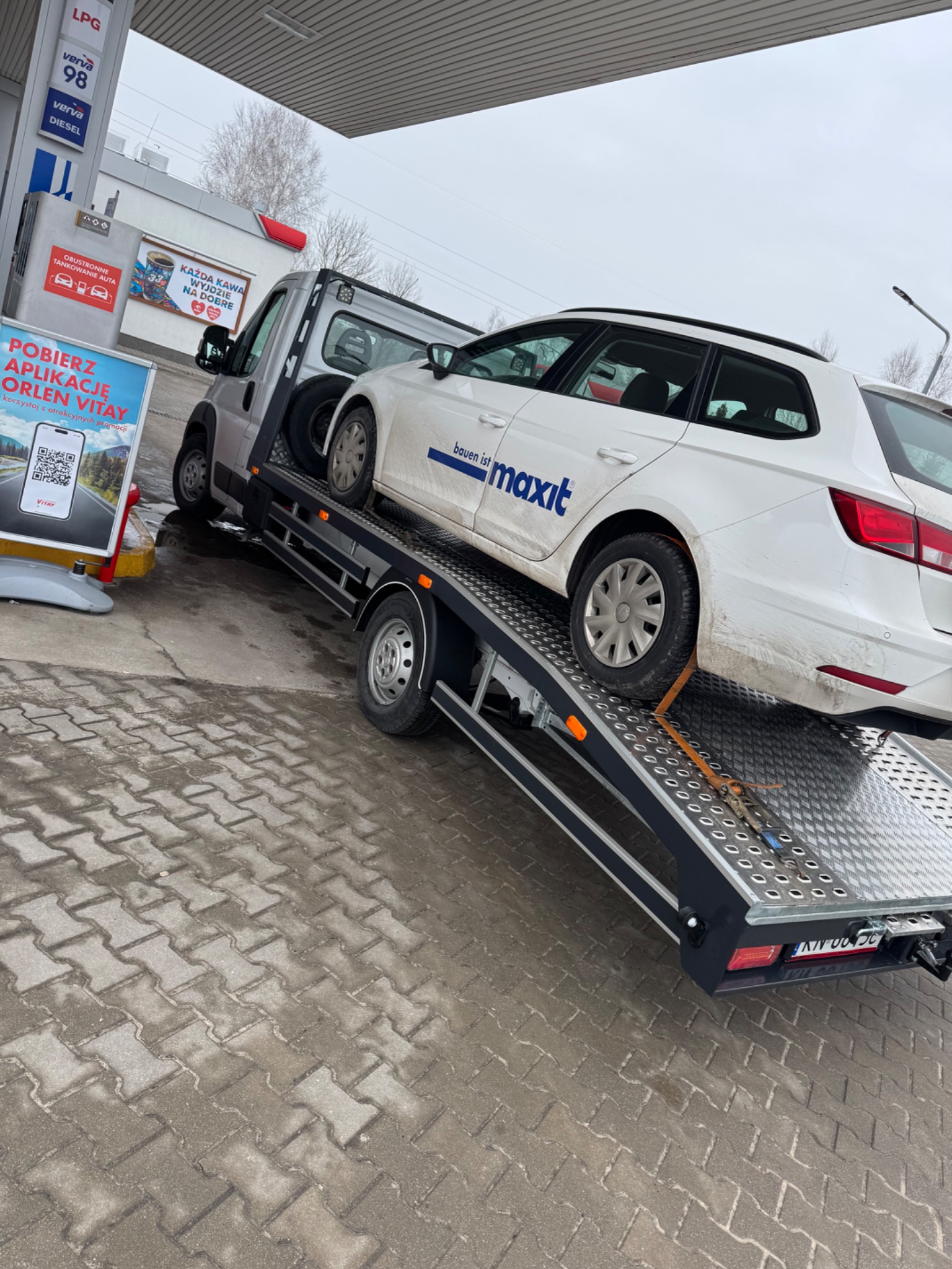 Laweta na stacji benzynowej przewozi biały samochód osobowy z napisem 'bauen ist maxit', zabezpieczony pasami transportowymi, widoczny dystrybutor LPG i reklama aplikacji Orlen Vitay.