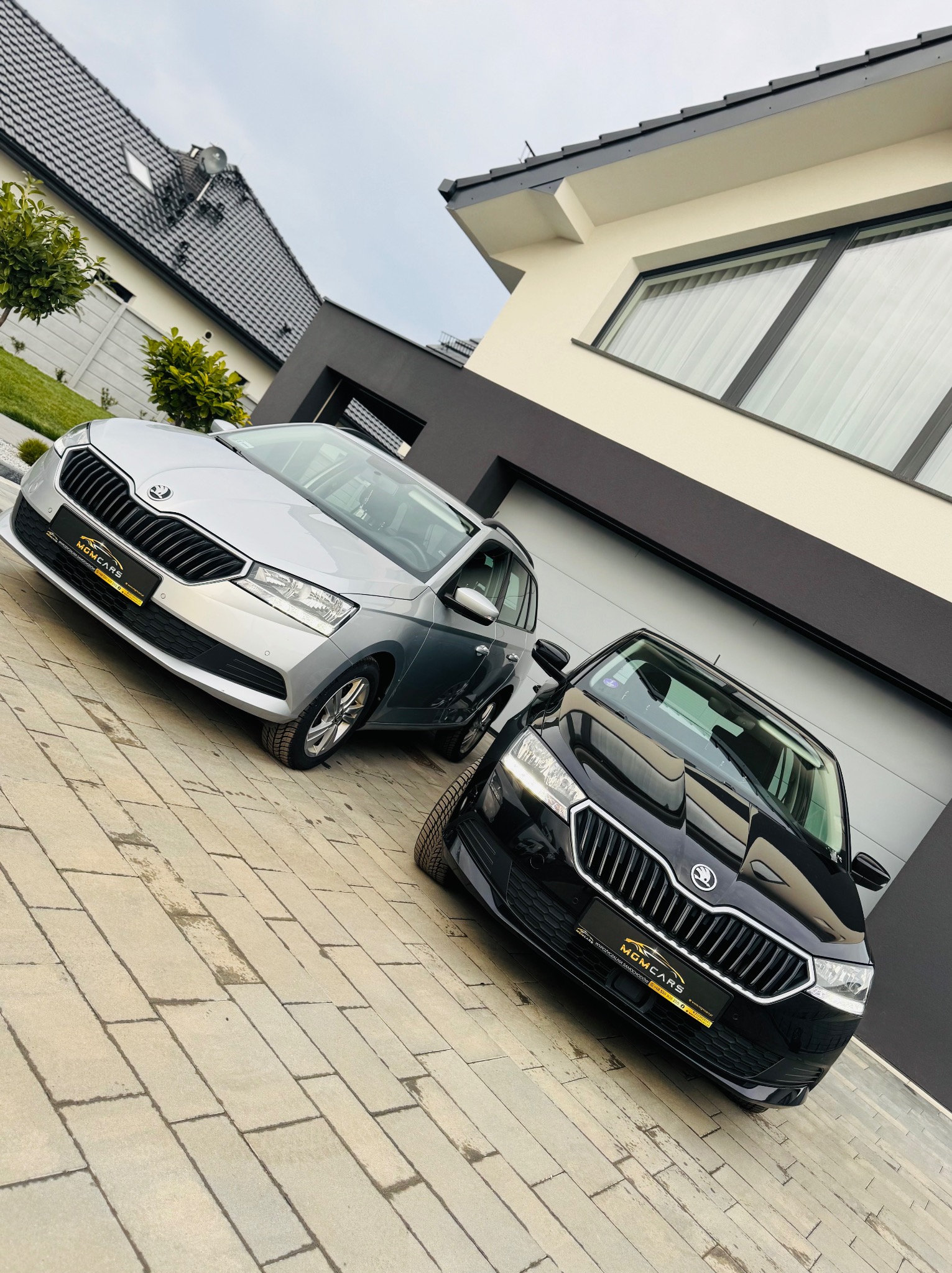 Dwa samochody Skoda, srebrny i czarny, stoją zaparkowane na podjeździe przed nowoczesnym domem z szarą bramą garażową.