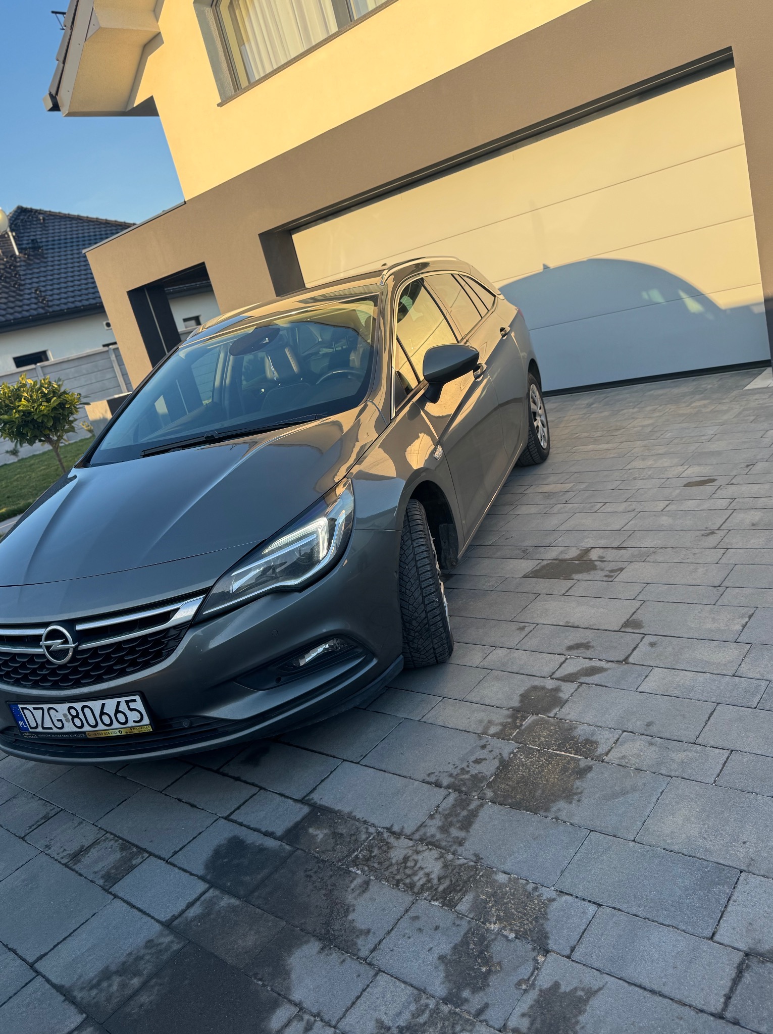 Szary samochód kombi marki Opel Astra zaparkowany na posesji przed nowoczesnym domem z garażem, widoczne ślady wilgoci na kostce brukowej.