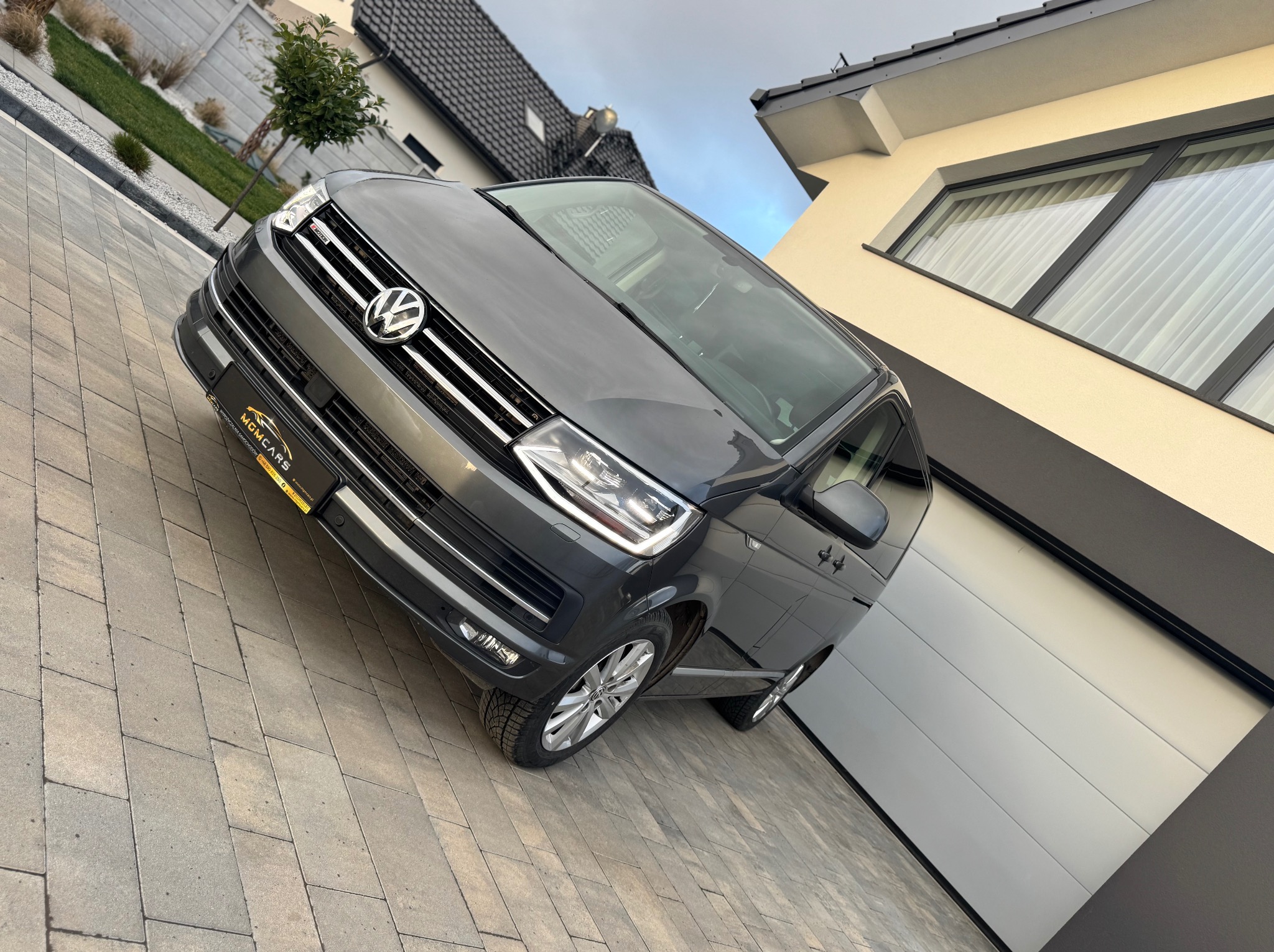 Szary Volkswagen Transporter T6 na brukowanym podjeździe przed nowoczesnym domem z białą bramą garażową i zadbanym trawnikiem.