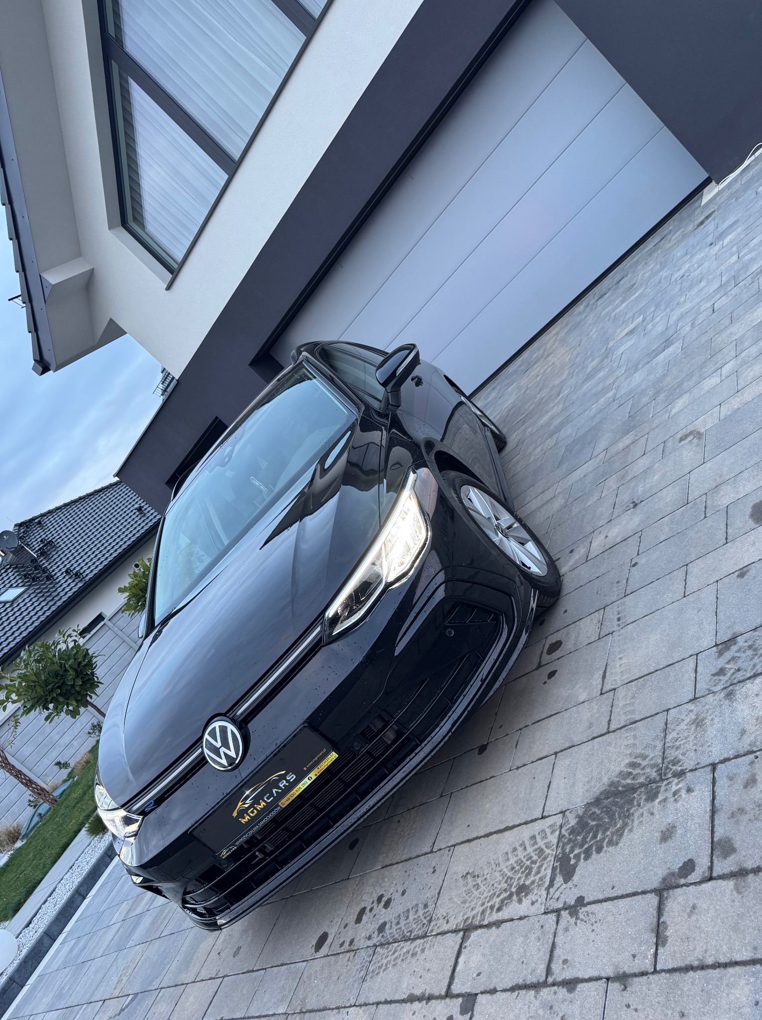 Czarny samochód osobowy marki Volkswagen zaparkowany na kostce brukowej przed nowoczesnym domem z szarą bramą garażową i dużym oknem.