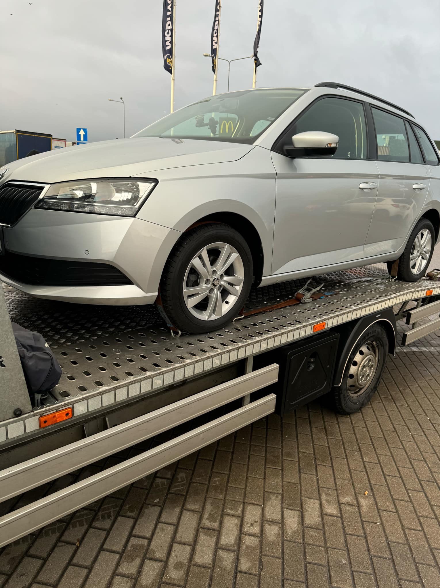 Srebrny samochód osobowy Skoda Fabia kombi zabezpieczony pasami transportowymi na platformie lawety, widok z boku, na tle pochmurnego nieba i flag z logo.