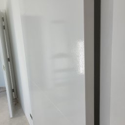 Remont click - Świeżo pomalowane na biało drzwi wewnętrzne z widocznymi zawiasami zabezpieczonymi taśmą malarską, w tle fragment ściany i podłogi w trakcie remontu.