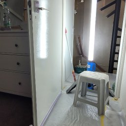 Remont click - Pokój w trakcie odświeżania: świeżo pomalowana na biało ściana z widocznym odbiciem światła, podłoga zabezpieczona folią malarską, wiadro z farbą i narzędziami malarskimi, drabina i lampa...