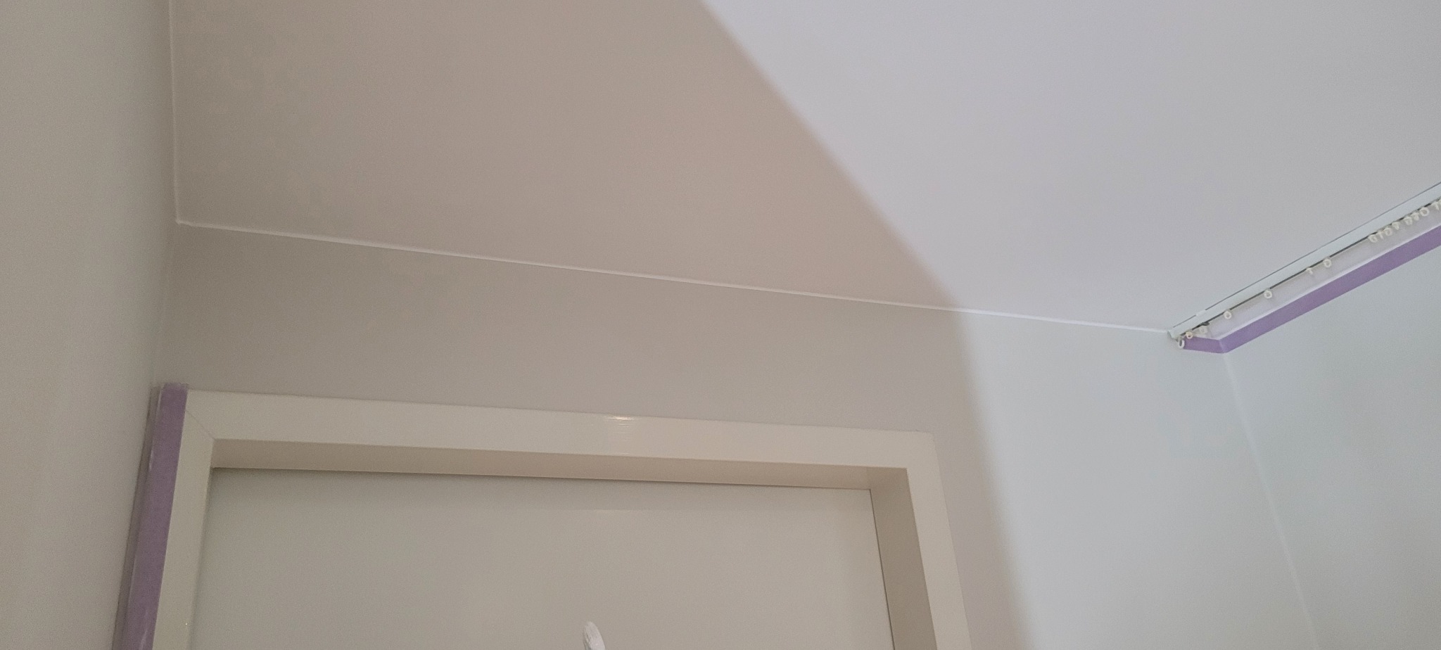 Świeżo pomalowany narożnik pokoju z listwą przypodłogową i karniszem, widoczne delikatne przejścia kolorów na ścianach i suficie, zarys framugi drzwi.