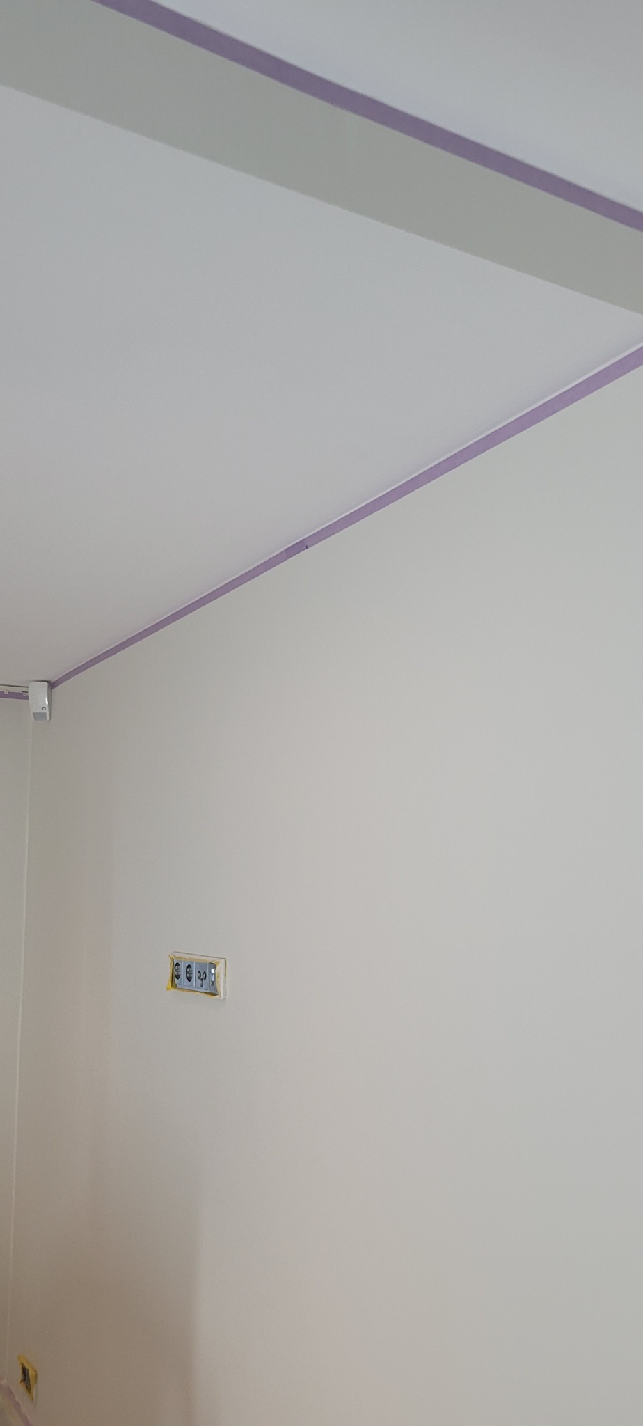 Świeżo pomalowana ściana i sufit w jasnym odcieniu, zabezpieczone fioletową taśmą malarską przy krawędziach, z widocznymi gniazdkami elektrycznymi zabezpieczonymi taśmą.